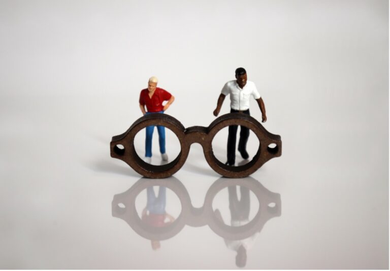 Two figures look through a giant pair of glasses and consider their reflection on the shiny floor.